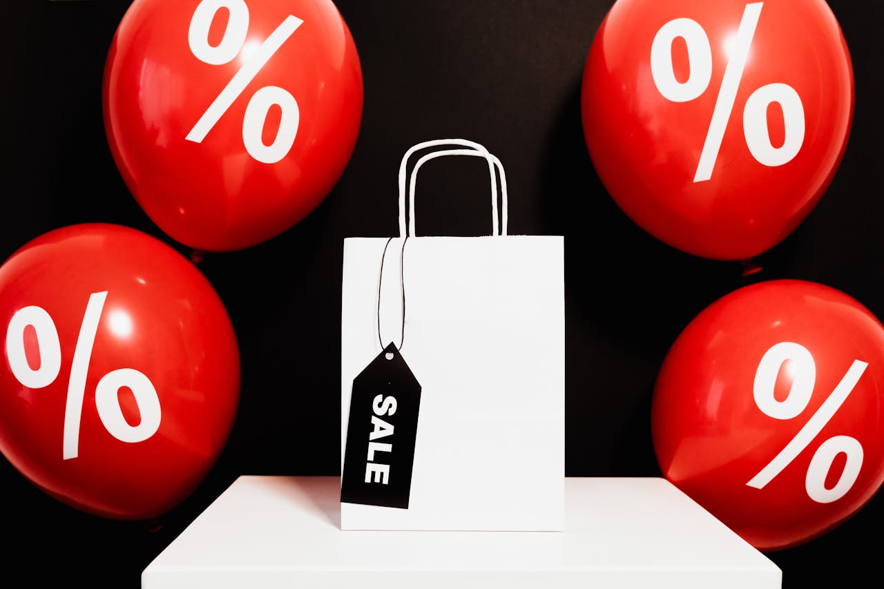 White shopping bag with sale tag surrounded by red balloons displaying percentage signs.